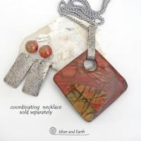These sterling silver earrings have a modern, earthy organic flair. They feature handcrafted sterling silver rectangles with a rustic hammered texture accented with natural red creek jasper stones. The stones have a beautiful natural pattern in earthy hues of mossy greens, brick red and orange. They have a coordinating necklace which is sold separately. 